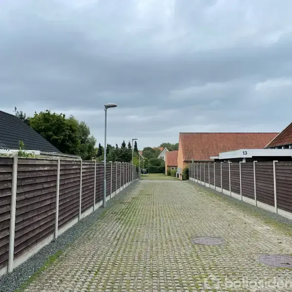 En smal brostensgade med høje træhegn på begge sider strækker sig mod horisonten. Omkringvejen er præget af et par ensformige, lavtliggende huse med røde tage under en overskyet himmel.