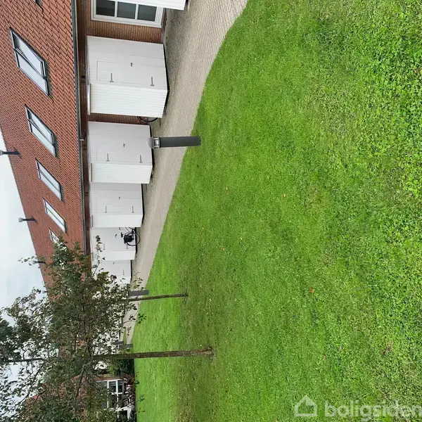 Rækkehusfacade med rødt tag, hvide døre og flere vinduer. Græsplæne og træer i forgrunden. Asfalteret sti løber langs huset, omgivet af en grøn have.