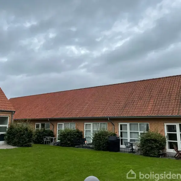 Bygning med rødt tegltag og vinduer, omgivet af en velplejet græsplæne og buske, under en overskyet himmel.