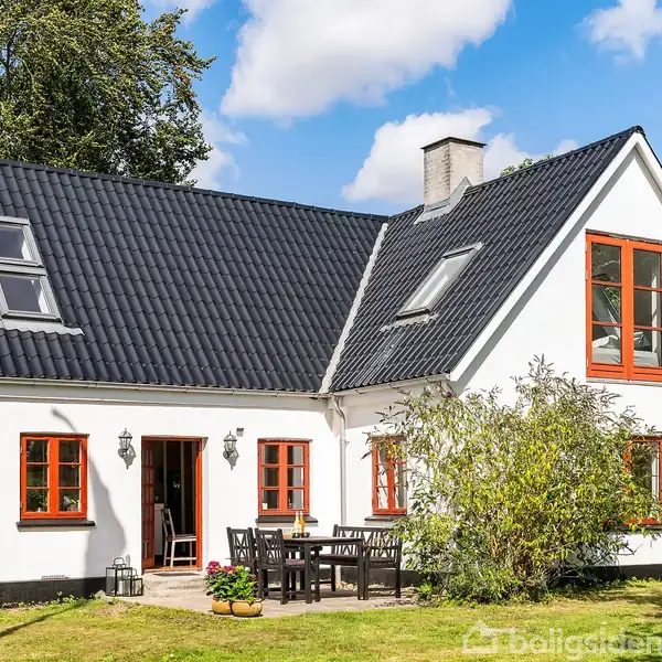 Et hvidt hus med sorte tagsten og røde vinduesrammer står i en grøn have. Et bord med stole ses foran huset, omkranset af buske og træer under en blå himmel.
