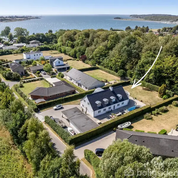 Et hvidt hus med sort tag står blandt andre huse i et grønt boligområde. En hvid pil peger på huset, og i baggrunden ses en kystlinje og havet.