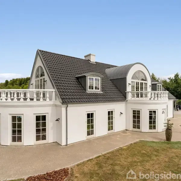 En hvid, to-etagers villa med gråt tegltag står stille. Villaen har store vinduespartier og en balkon. Den ligger i en omgivet af grønne buske og træer på en solrig dag.