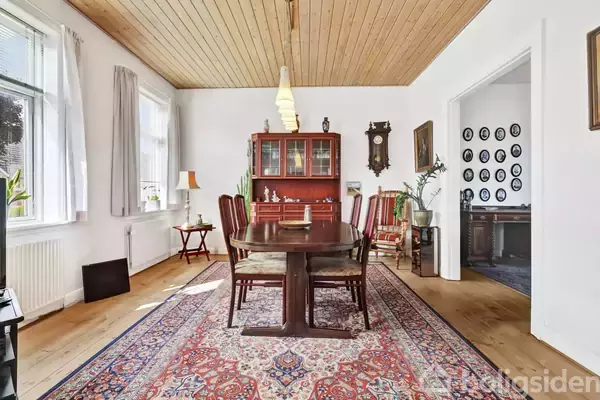 Et spisebord med stole står på et orientalsk tæppe i et lyst rum med trægulve og hvidmalede vægge. En skænk, ur og billeder hænger på væggene.