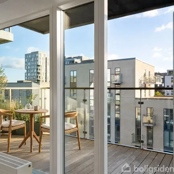Balkon med to træstole og et rundt bord. Står stille i moderne bymiljø med lejlighedsbygninger. Sollys skinner ind gennem glasdøre.