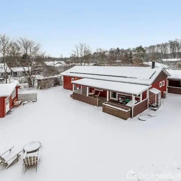 Rødt hus med overdækket terrasse står i snedækket haveomgivelser. Et mindre udhus ses til venstre, og træer omkranser området i baggrunden. Bord og stole dækket af sne.
