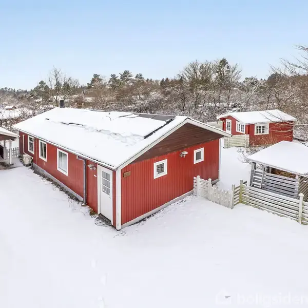 Rødt hus står stille i et snedækket landskab med tilhørende mindre bygninger og terrasse. Hvide havemøbler er delvist dækket af sne, omgivet af spredte træer.