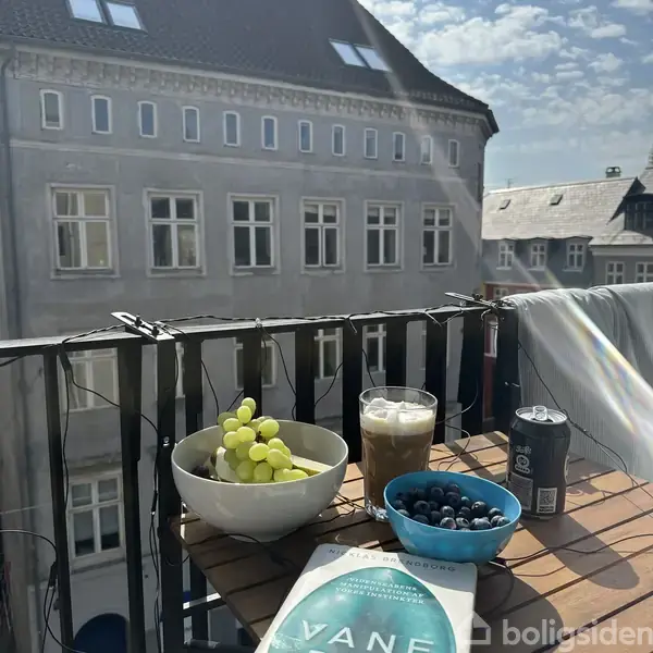 Både med grønne druer, en glas iskaffe og en dåse øl står på et bord på en altan. En skål med blåbær er også til stede. Byhus ses i baggrunden.