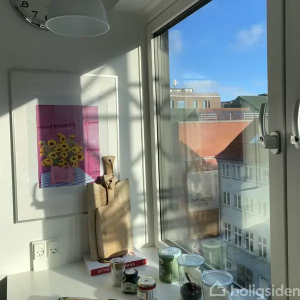 Et letoplyst køkkenhjørne med en dekoreret væg indeholder et kunstværk af solsikker i en lyserød vase. To skærebrætter står ved siden af. Udsigt til byhuse ses gennem vinduet.