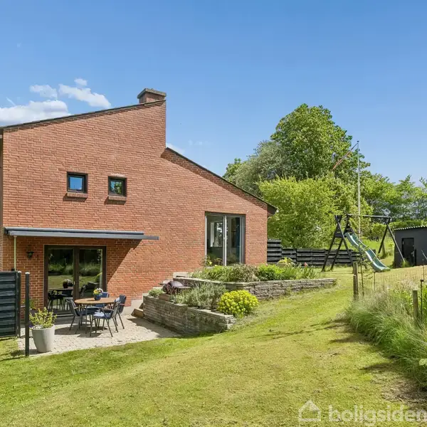 Rødt murstenshus med terrasse møbleret med bord og stole. Huset står i en grøn have med græsplæne og legeredskaber, omgivet af træer og buske under en klar blå himmel.