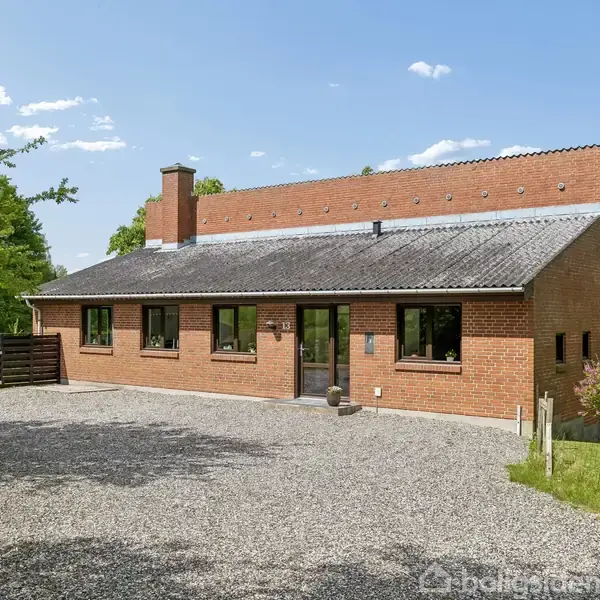 Et murstenshus med fladt tag står stille foran en grusvej, omgivet af grønne træer og en mark. Sollys skinner på huset under en næsten skyfri himmel.