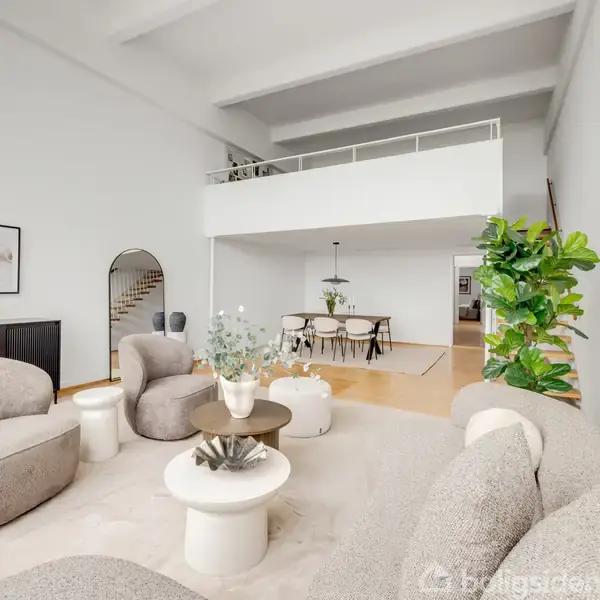 Stue med moderne møblering; komfortable lysegrå sofaer og stole omkring et sofabord. Spisebord med stole i baggrunden under en hems. Rummet er lyst med store planter og minimalistisk dekoration.