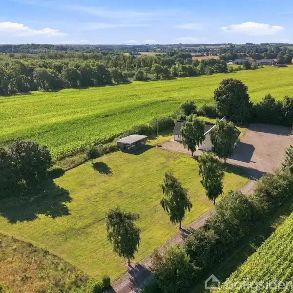 En grøn mark med en lille vej og enkelte træer omkranser en åben plads med en lille bygning. Landskabet er landligt med vidtstrakte marker i baggrunden under en blå himmel.