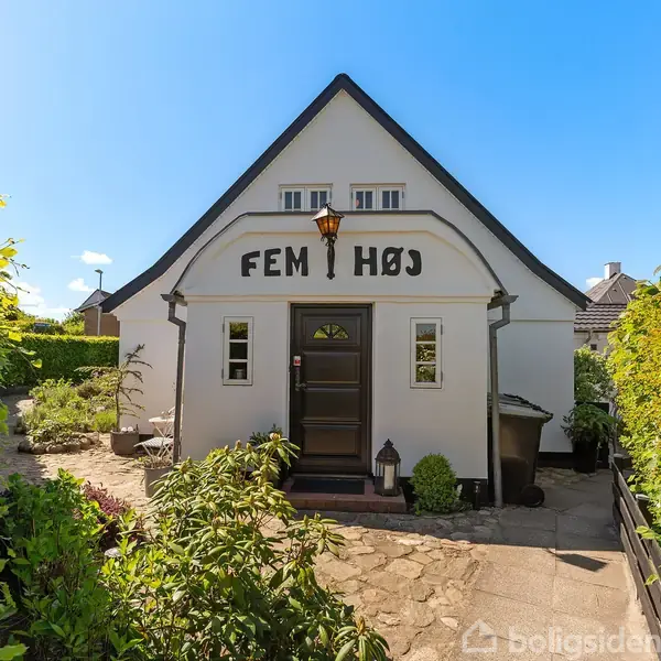 Hvidt, kompakt hus med skiltet "Fem Høj" over døren. Står foran en velplejet have med stenbelagt sti. Solrig dag med blå himmel.