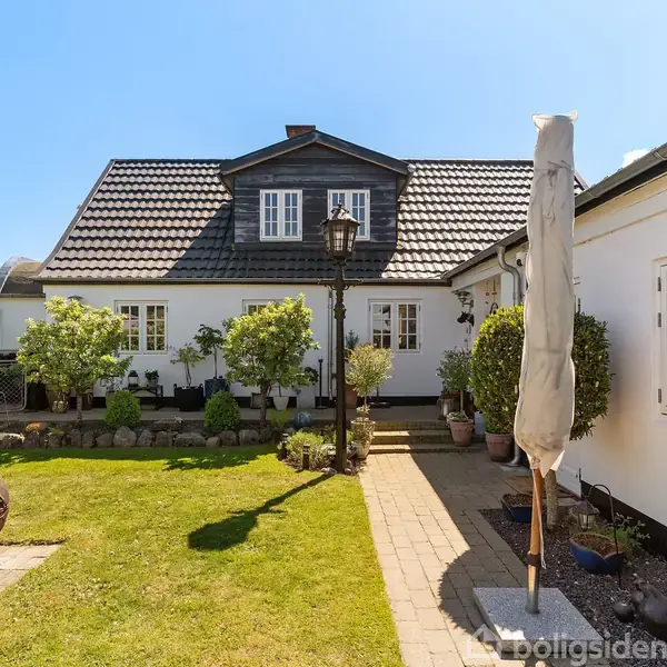 Et hvidt hus med sorte tagsten står i en velplejet have. Et rustikt ildsted og en sammenfoldet parasol ses i forgrunden, omringet af grønne buske og træer.