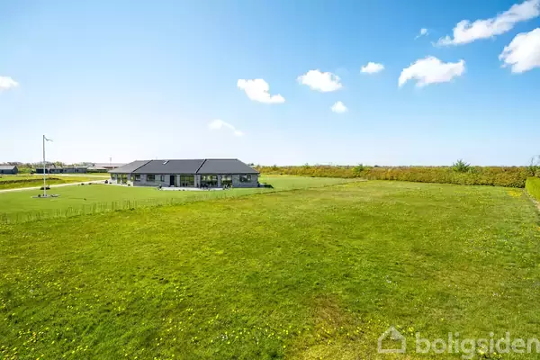 Hus står på en stor grøn græsplæne med en flagstang ved siden af, under en blå himmel med få skyer. Grunden er delvis indhegnet med hække.