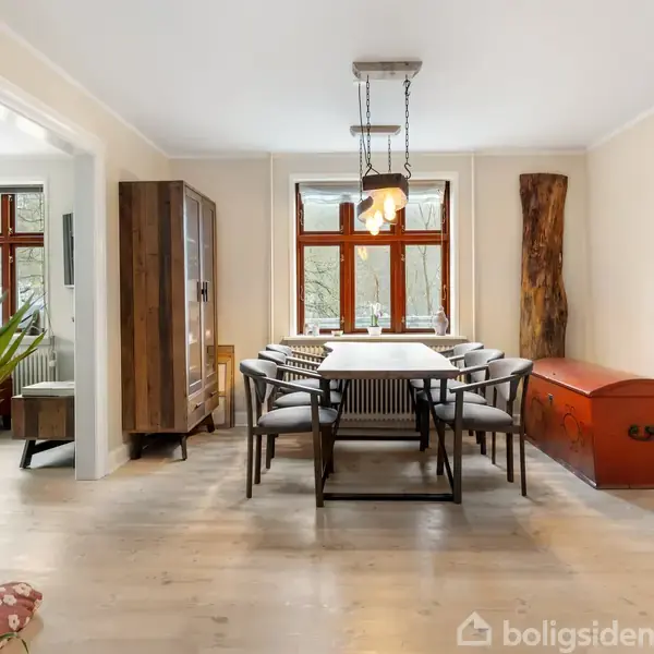 En spisestue med et stort bord og otte stole, omgivet af hvide vægge og store vinduer, en trækommode til venstre og en rød kiste i hjørnet.