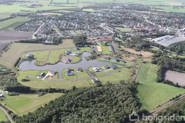 Landskabet viser et luftfoto over en grøn landlig by med huse omkring kunstige søer. Området er omkranset af skove og marker, med bebyggelse i baggrunden.