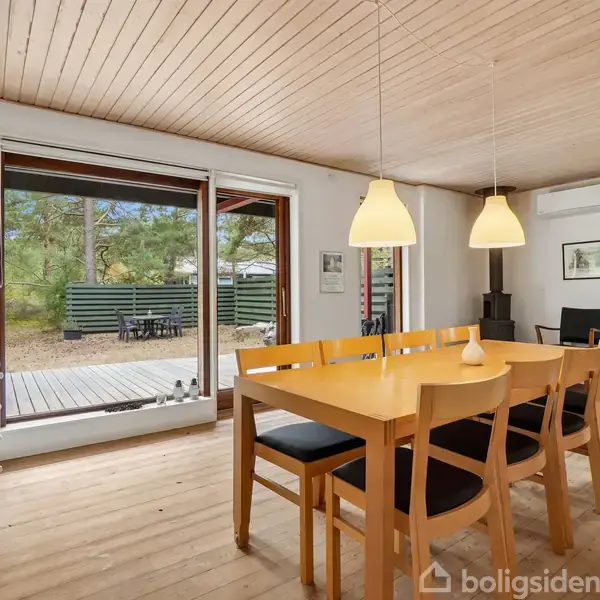 Spisebord med seks stole står midt i et lyst rum med trægulv og træloft. Store vinduer og en skydedør åbner mod en terrasse og have. To hængelamper over bordet.