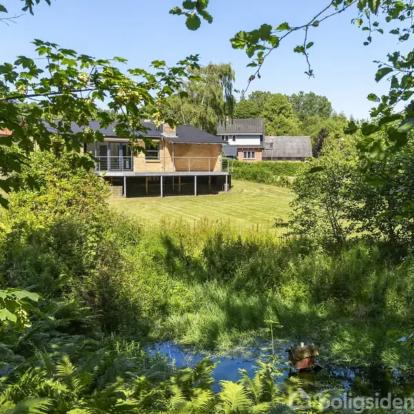 Et moderne hus ligger på en åben græsplæne omgivet af frodig skov. I forgrunden ses en lille dam og tæt bevoksning, hvilket skaber en naturskøn og privat atmosfære.