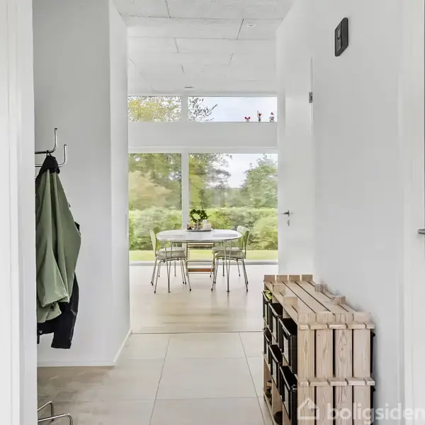 Entreen fører til et lyst rum med et rundt spisebord og stole foran store vinduer, der viser en udsigt til en grøn have. En frakke hænger ved døren.