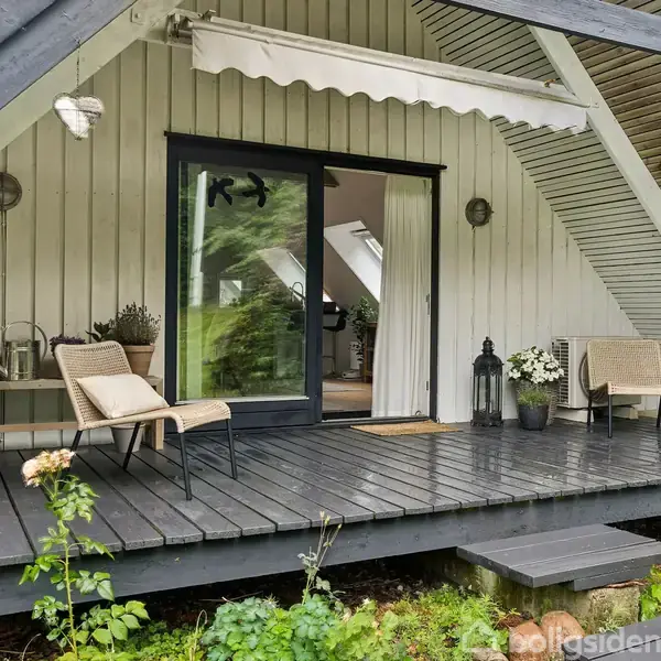 A-husets terrasse har to stole og et bord på mørkt trædæk. En grill står til venstre, planters i krukker til højre. Indersiden ses gennem glasdøre under en markise.