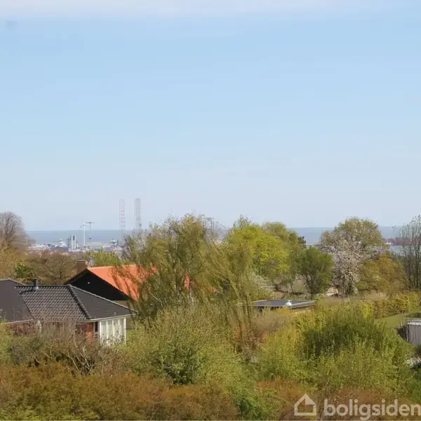 Hus står stille i forgrunden omgivet af grønne træer og buske, med udsigt over bylandskab og hav i baggrunden under en klar blå himmel.