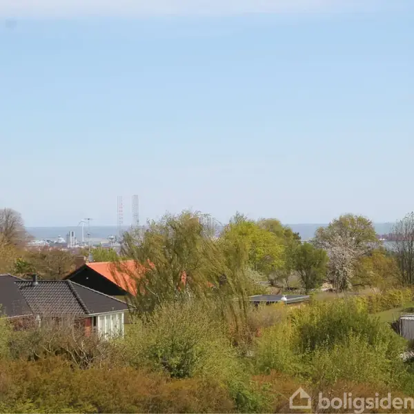 Huse ligger stille blandt frodige træer under en blå himmel, med hav og en by i det fjerne baggrund, giver visuelt perspektiv til landskabet.