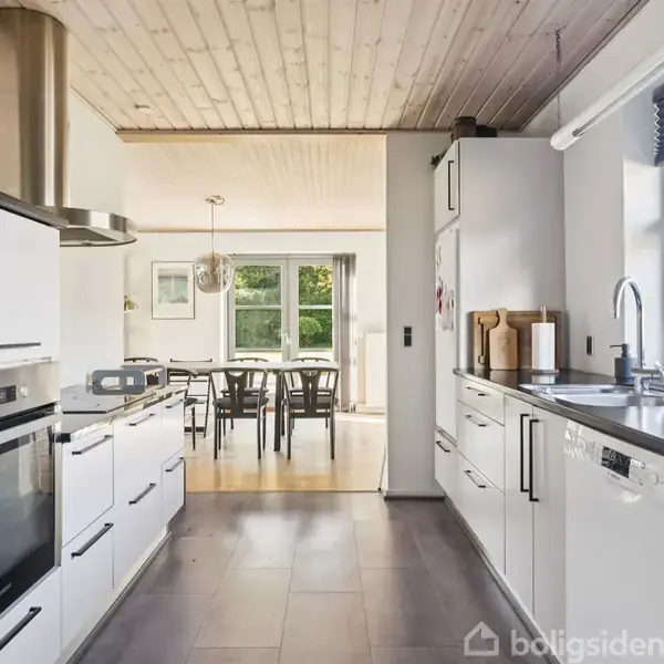 Et moderne køkken med hvide skabe og sorte bordplader. Der er en ovn til venstre og en vask til højre. Foran er der et spisebord med stole i en lys spisestue.