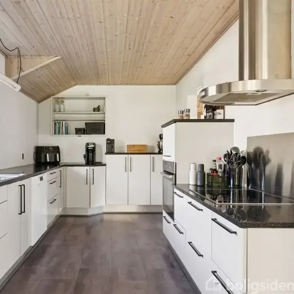 Moderne køkken med hvide skabe og sorte bordplader. Køkkenmaskiner og krydderier er placeret på bordet. Store vinduer giver naturligt lys. Lys trægulv og loft skaber en varm atmosfære.