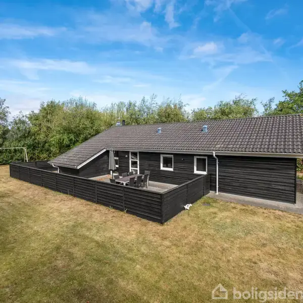 Sort træhus står stille i en åben have, omgivet af grønne buske og træer. Der er en terrasse med siddemøbler og en fodboldmål i baggrunden.