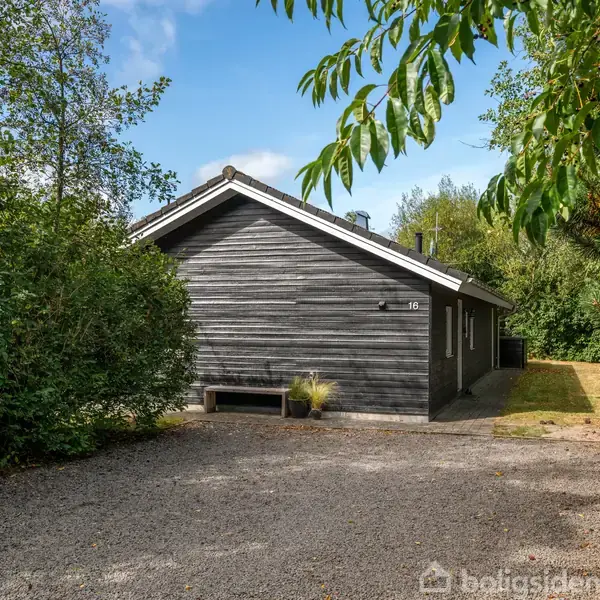 Et gråt træhus står stille omgivet af grønne træer og buske. En lille grussti fører op til huset, der har en hvid tagkant. Huset nummer "16" vises ved indgangen.