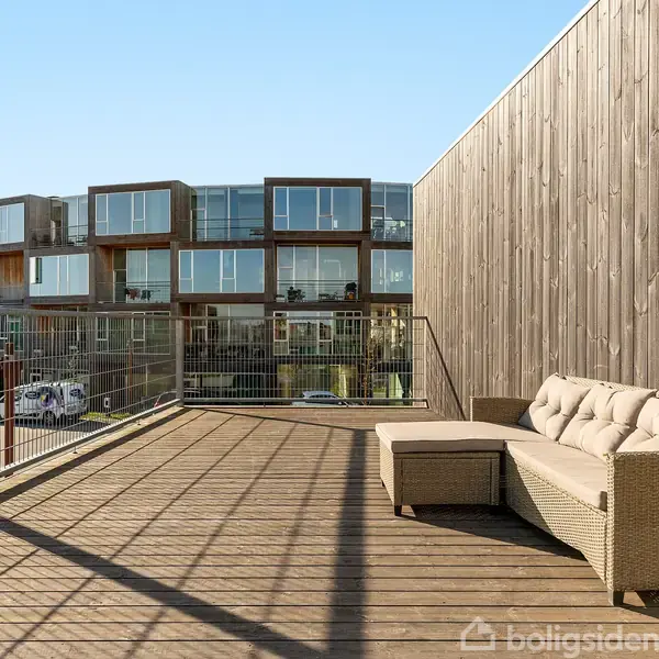 En beige sofa står på en stor træterrasse med udsigt over moderne lejlighedsbygninger. Området er solrigt og omgivet af klare, åbne omgivelser.