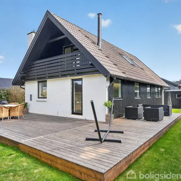 Hus med skråt tag står stille på stor træterrasse med havemøbler. Græsplæne og hegn omgiver huset i et boligområde.