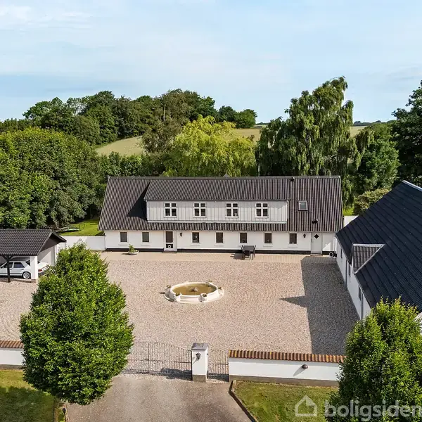 Et stort, hvidt hus med sort tag omkranser en grusbelagt gårdsplads med en rund fontæne i midten. Omgivet af træer og grønne bakker i baggrunden.