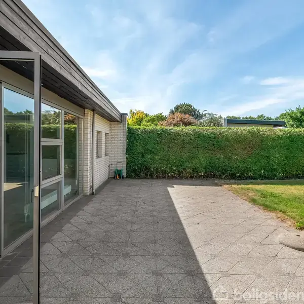 En flisebelagt terrasse med glasdør står åben mod en veltrimmet græsplæne, omgivet af en høj, grøn hæk under en klar blå himmel.