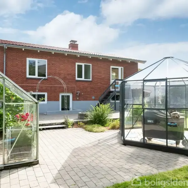 Rød murstenshus med to drivhuse i haven. Trapper fører til en terrasse, omgivet af fliser og græsplæne, under en blå himmel med lette skyer.
