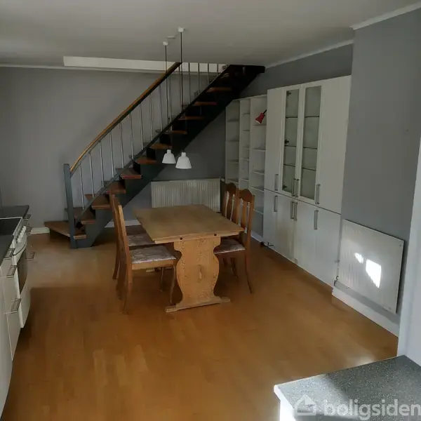 Træbord med fire stole står stille i et spisekøkken med trægulv. Baggrund indeholder en trappe, hvide skabe og grå vægge.