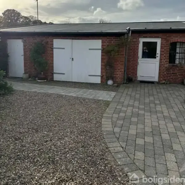 En murstensbygning med en hvid dør og garageport. Foran er der en belagt stenvej og stenbed. Området er bevokset med planter og små træer ved væggen.