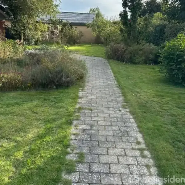 En flisebelagt sti fører gennem en grøn have med velplejet græs og tætte buske langs siderne under en klar himmel.