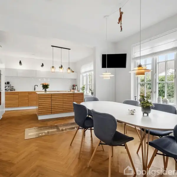 Et moderne køkken med hvidt bord og sorte stole står i et rum med trægulv og store vinduer. Køkkenet har træpaneler og hængelamper. En plante ses på bordet.