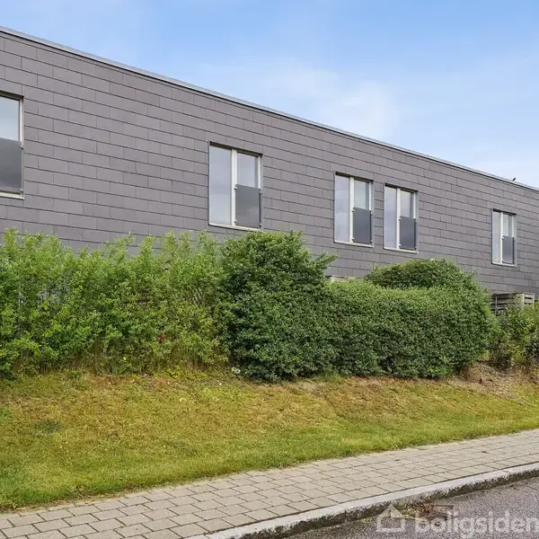 Bygning med grå facade og rektangulære vinduer står stille bag en hæk. Fortovet og en vej skaber en urban kontekst, mens en gadelampe ses i højre side.
