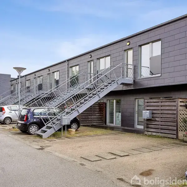 Rækkehuskompleks med udendørs metaltrapper fører til øverste etage. Parkerede biler langs vejen foran. Træhegn og mindre haveområder nær indgangen. Klar blå himmel i baggrunden.