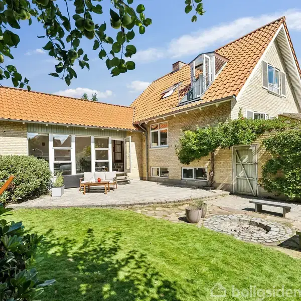Hus med rødt tegltag står i solrig have. Der er terrasse med havemøbler og grøn græsplæne omgivet af buske og træer, skaber en fredelig udendørs atmosfære.