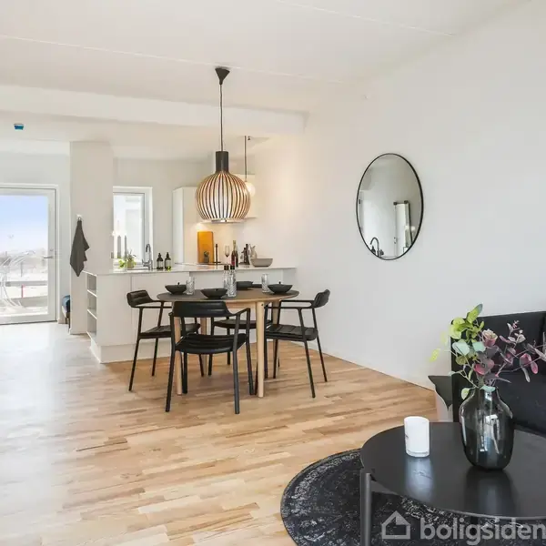 En spisestue med et sort spisebord og fire stole står midt i rummet. En rund lampe hænger over bordet. Til højre er en sort sofa med en rund lampe ved siden af.