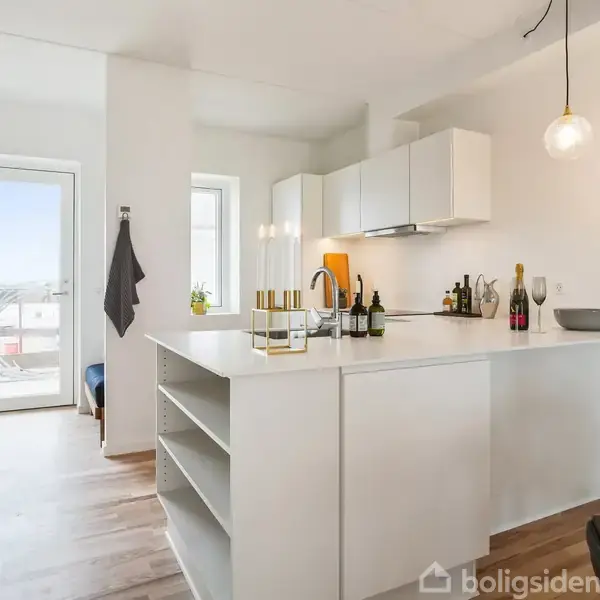 Køkken med hvidt køkkenbord, og indbyggede skabe, flasker og kogeplader. Rummet er lyst med trælaminatgulv og har en glasdør i baggrunden, der fører til en balkon eller veranda.