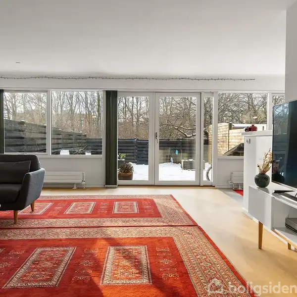 Stue med en stor fladskærms-tv på hvidt møbel, røde tæpper på trægulv, en grå lænestol, og glasdøre der fører ud til en snedækket terrasse og skov.