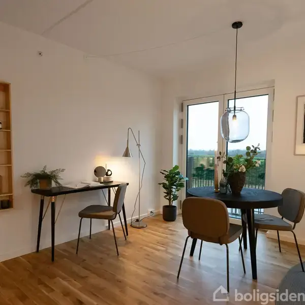 Et lyst rum med trægulv indeholder et lille rundt spisebord med tre stole, en skrivebordslampe på et skrivebord, hylder med små genstande og en stor plante ved et vindue.