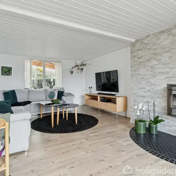 En stue med en lys grå sofa arrangeret rundt om et sort rundt tæppe. Et sofabord står foran sofaen. Et tv er placeret på et træmøbel. Der er en pejs indbygget i væggen, mens gulvet er af lyse træplanker.