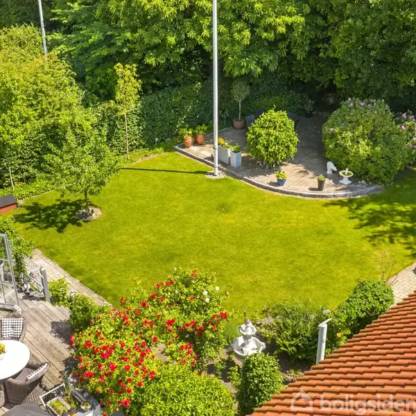 Cirkulær terrasse med havemøbler grænser op til en velplejet græsplæne omgivet af tætte træer og buske. Solbelyst have fremhæver blomstrende planter og spredte dekorative elementer.