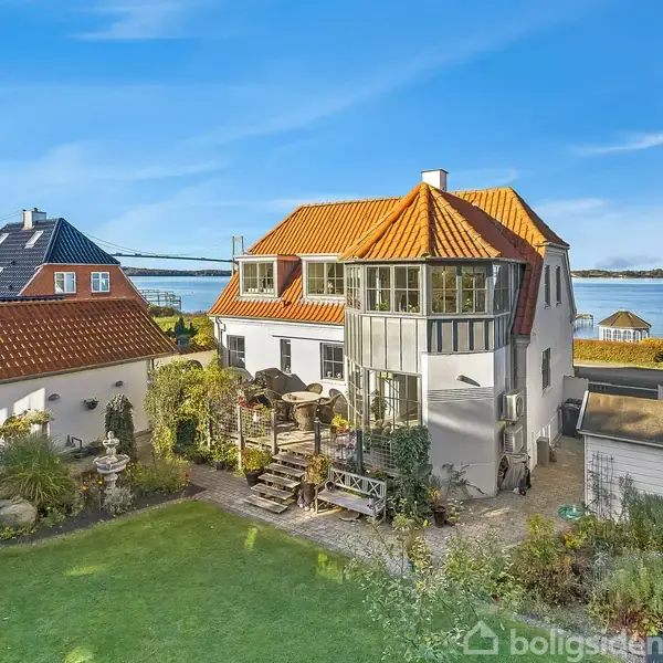 Huset står i en frodig have med udsigt over vandet. Det har en rød tegltag, flere vinduer og en terrasse med havemøbler, omgivet af øvrige huse og grønne omgivelser.